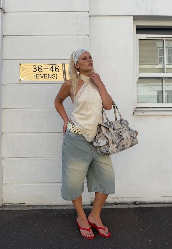 Žena v krémovom halter top-e, šortkách po kolená, červených sandáloch a šatke na hlave stojí pri bielej stene s tabuľou s číslom domu a drží veľkú vzorovanú kabelku.