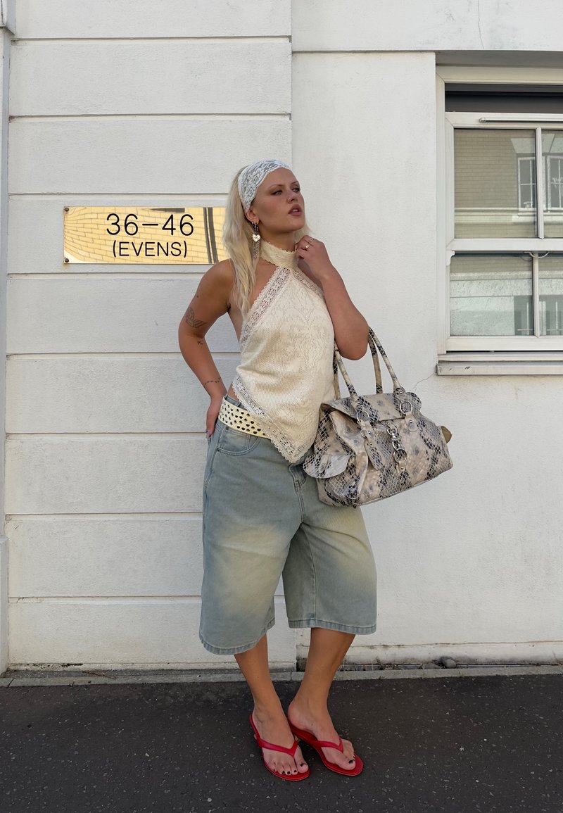 Femme en haut dos nu couleur crème, short jusqu’aux genoux, sandales rouges et foulard sur la tête, debout près d’un mur blanc avec une plaque indiquant le numéro de la maison, tenant un grand sac à main à motifs.
