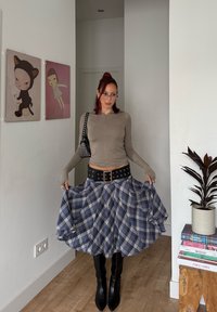 Maglia marrone a maniche lunghe, gonna a quadri blu e bianca, cintura larga nera con borchie d'argento, stivali neri a punta al ginocchio, ambiente interno.