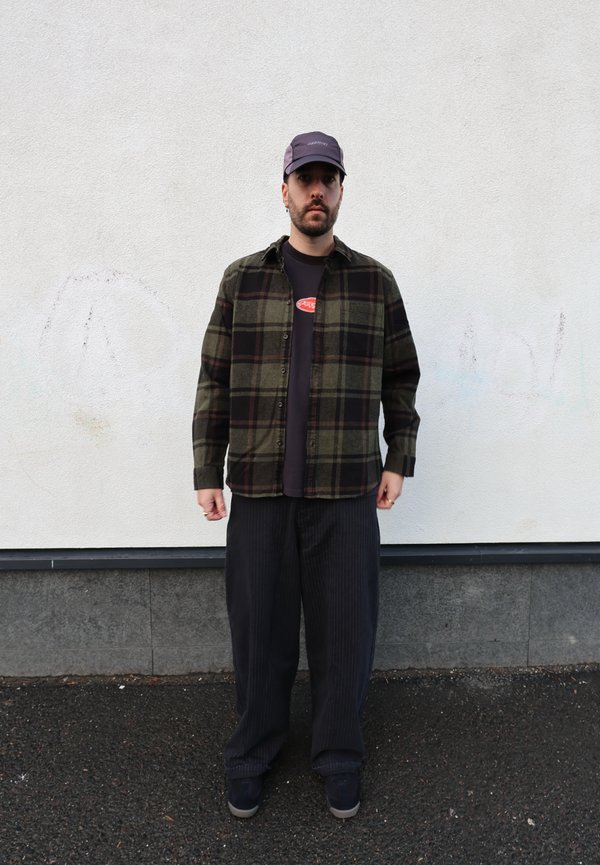 Koszula flanelowa w kratę w kolorach zielonym, czarnym i czerwonym, noszona na czarnej koszulce z czerwonym logo. Szerokie spodnie w ciemne paski i szare trampki.