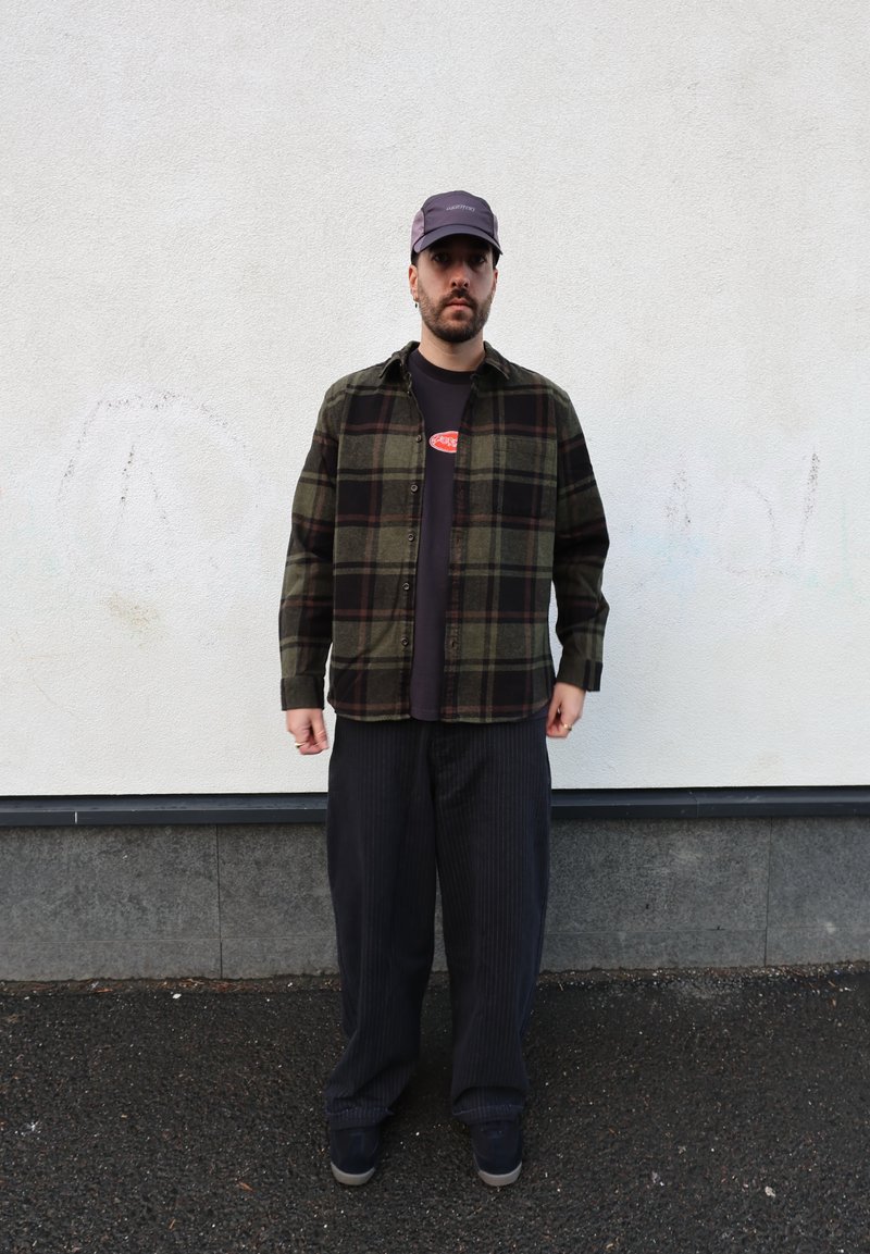 Karirana flanelasta srajca v zeleni, črni in rdeči barvi, nošena čez črno majico z rdečim logotipom. Široke hlače s temnimi črtami in sivi supergi.