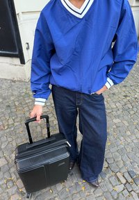 Veste légère bleue avec des rayures blanches, associée à un jean large et foncé. Une valise à roulettes noire présente une surface texturée et une fermeture éclair.