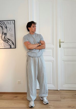Graues Kurzarm-T-Shirt und passende lockere Sweatpants, kombiniert mit grauen und weißen Turnschuhen, vor einer neutralen Wand mit einem eingerahmten Poster.