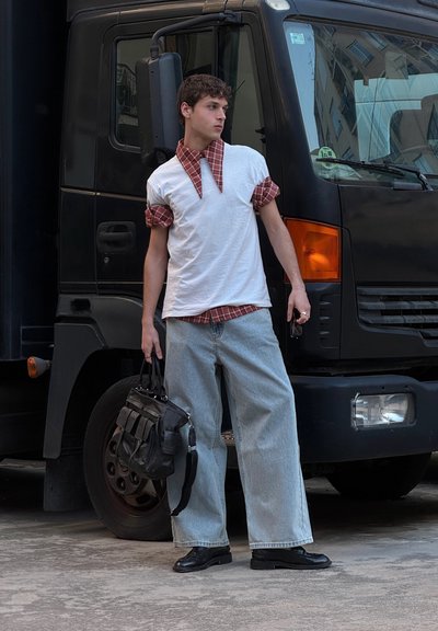 Camiseta blanca con un gran cuello de cuadros rojos, jeans de pierna ancha en azul claro, zapatos negros y un bolso negro. El fondo presenta un camión aparcado.
