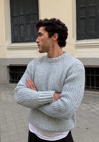 Maglione grigio chiaro a coste con scollo rotondo e polsini ampi, indossato sopra una camicia bianca, caratterizzato da una maglia strutturata e una vestibilità comoda.