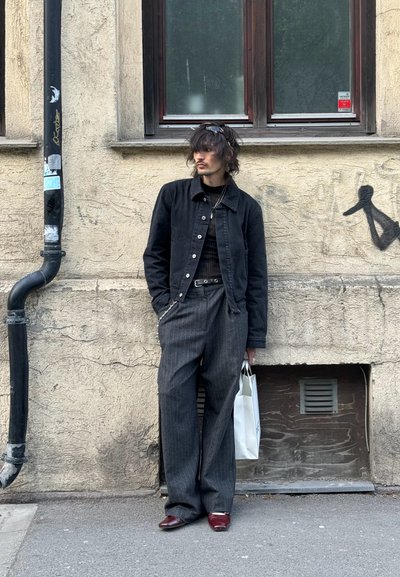 Chaqueta de mezclilla negra sobre una blusa negra de canalé, pantalones de pierna ancha a rayas grises y zapatos burdeos. Sostiene un bolso blanco; tiene el cabello desordenado.