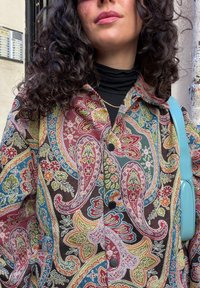 Femme aux cheveux bouclés portant une veste à motif cachemire multicolore, un col roulé noir, une chaîne en or, et un sac bleu clair porté sur l'épaule.