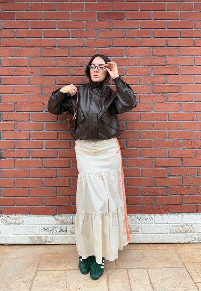 Mujer de pie contra una pared de ladrillo rojo, lleva una chaqueta de cuero negra, falda blanca con volantes y rayas naranjas, zapatillas verdes y gafas.