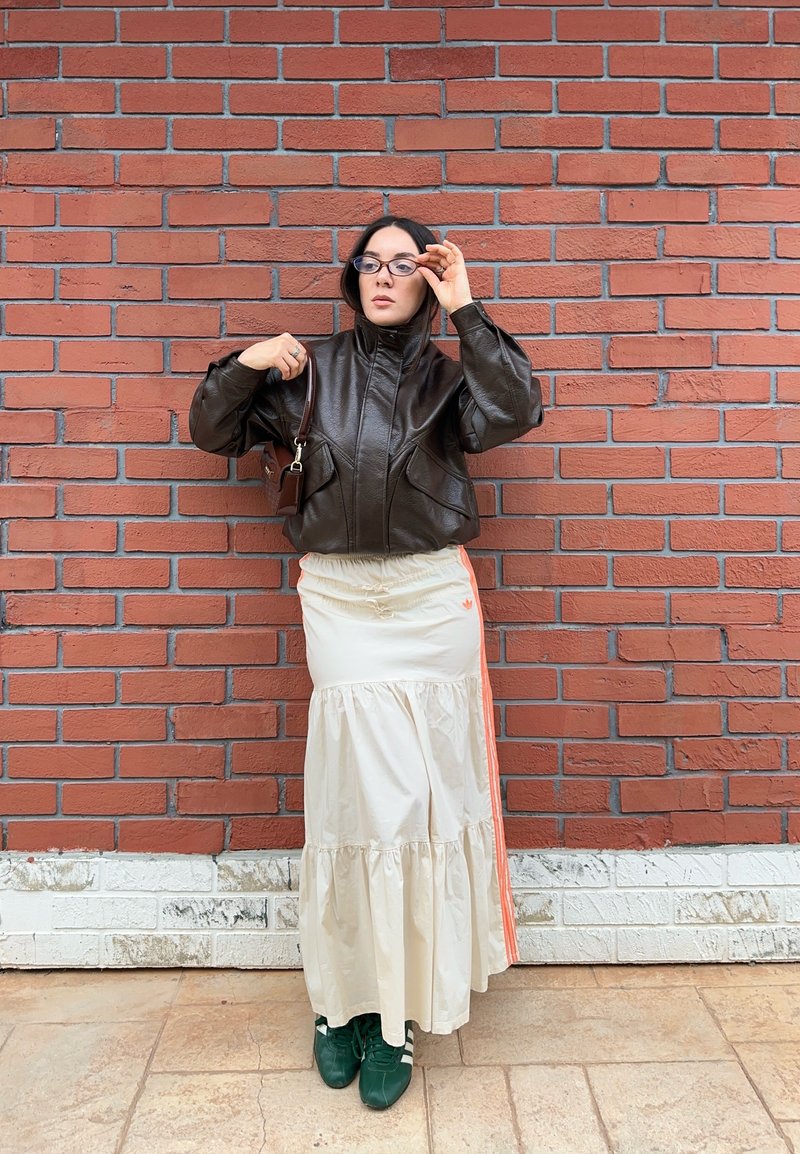 Sieviete stāv pie sarkanā ķieģeļu sienas, valkājot melnu ādas jaku, baltu vairākslāņu svārku ar oranžām svītrām, zaļas sporta kurpes un brilles.