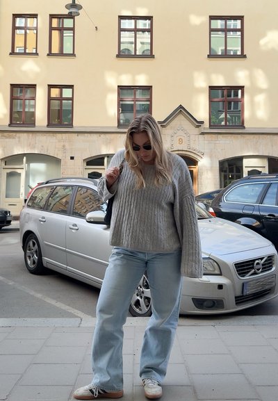 Suéter de punto trenzado gris, jeans acampanados azul claro y zapatillas. Gafas de sol puestas; entorno urbano con coches aparcados y edificios de fondo.