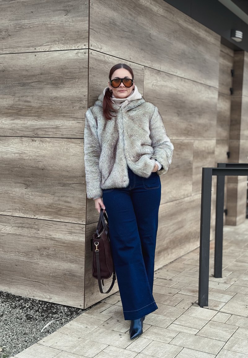 Jacke aus Kunstpelz in Hellgrau, kombiniert mit dunkelblauen Jeans mit weitem Bein. Schwarze Lederstiefel mit spitzer Spitze und eine dunkle Tasche mit Schulterriemen.