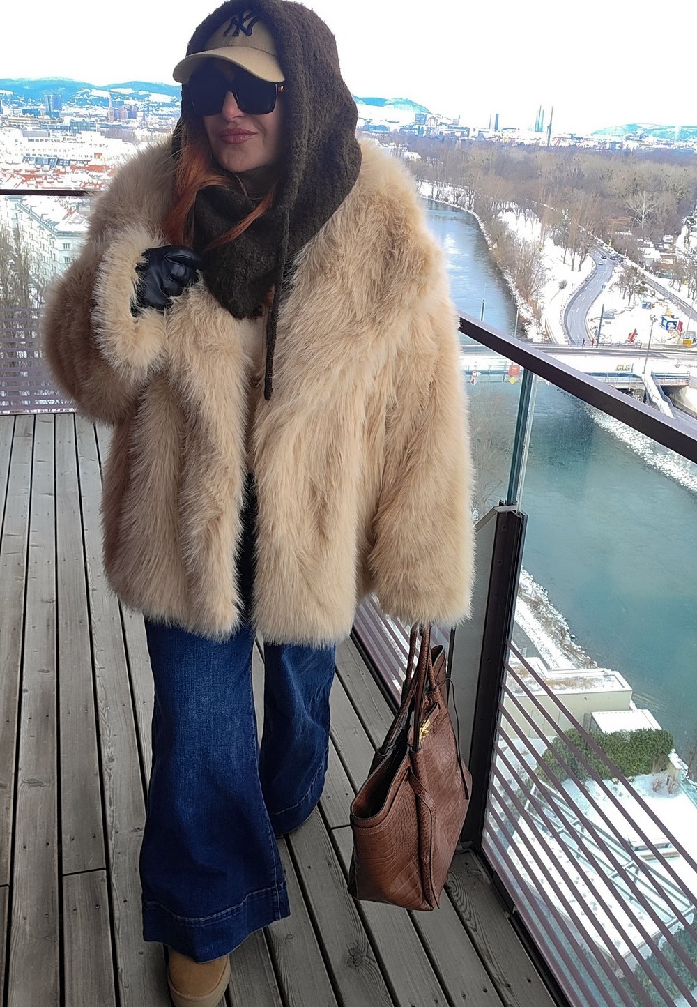 Personne portant un manteau en fourrure beige, une écharpe foncée, des lunettes de soleil et une casquette, tenant un sac à main marron sur un balcon donnant sur une rivière et un paysage urbain enneigé.
