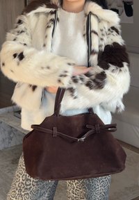 Borsa in suede marrone con due manici e un accento a fiocco decorativo. Indossata con un maglione bianco lavorato a maglia e pantaloni con fantasia. Cappotto in pelliccia sintetica sullo sfondo.
