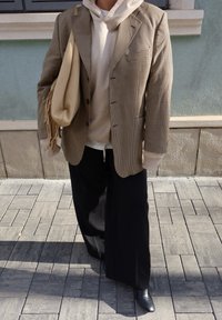 Oseba, oblečena v bež kapucar pod karo blazerjem, črne široke hlače, črni čevlji in nosi bež torbico z resami čez ramo na tlakovani pločnik.