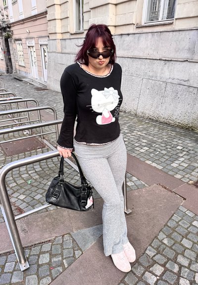 Camisa negra de manga larga con un gráfico blanco de Hello Kitty, pantalones acampanados grises, zapatillas rosas y un bolso negro. Entorno urbano al aire libre.
