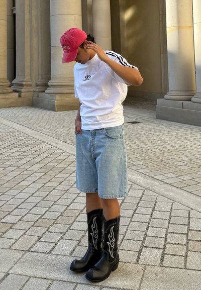 Camisa blanca de manga corta con logo negro, pantalones cortos de mezclilla azul claro y botas de cowboy negras con patrón de llamas blancas, de pie sobre adoquines.