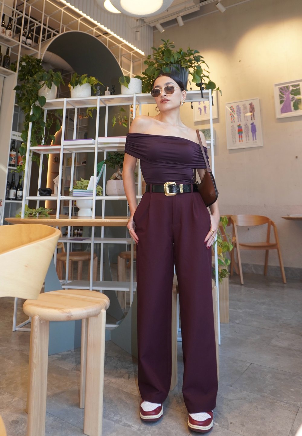 Burgundská off-shoulder halenka spojená s kalhotami s volnými nohavicemi, černým páskem a masivními teniskami. Sluneční brýle a minimální doplňky dotvářejí vzhled.