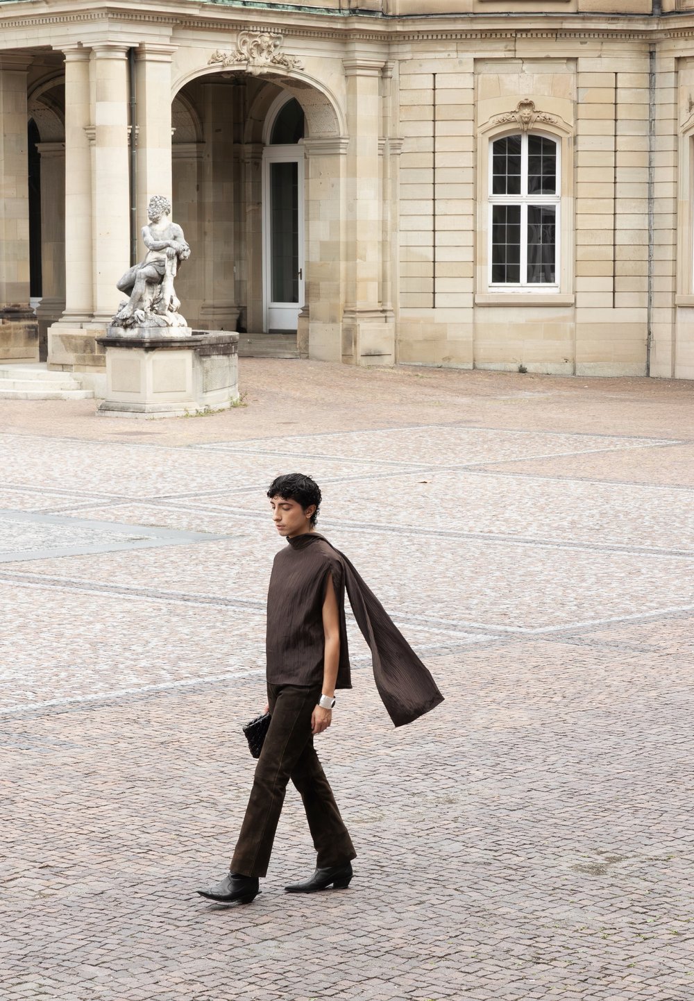 Persoon die over een kinderkopjesplein loopt, gekleed in een donkerbruin outfit met lange, vloeiende mouw, nabij een klassiek stenen gebouw en een standbeeld.