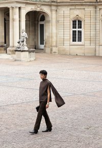Persoon die over een kinderkopjesplein loopt, gekleed in een donkerbruin outfit met lange, vloeiende mouw, nabij een klassiek stenen gebouw en een standbeeld.