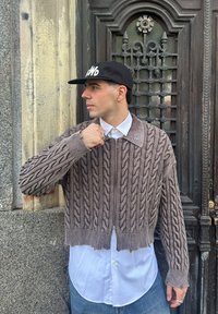 Camisola castanha de malha com padrão trançado, design cropped e fecho de correr à frente, usada sobre uma camisa branca com botões. Boné preto com logótipo branco.