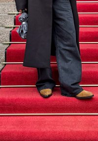 Brūni zamšādas apavi ar melnu zoli, apvienoti ar tumšiem džinsa biksēm, stāvot uz sarkanu paklāju kāpnēm ar zelta metāla margām.