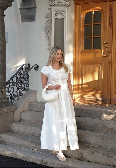 Vestido largo blanco de botones en la parte delantera, con mangas cortas y cuello. Bolsa de tejido y zapatos texturizados; de pie en unos escalones de piedra junto a una puerta de madera.