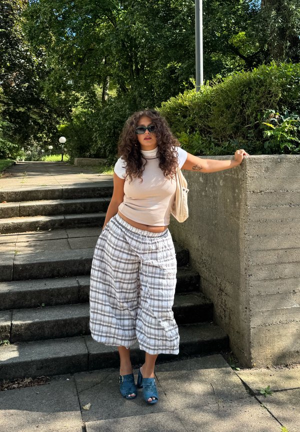 Femme aux cheveux bouclés et lunettes de soleil debout sur des marches extérieures, portant un haut sans manches, un pantalon large à carreaux et des chaussures à talons bleues.