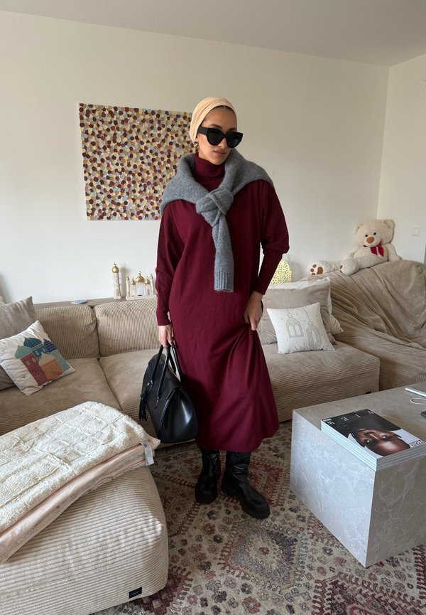 Mujer con un vestido granate, suéter gris sobre los hombros, botas y bolso negros, llevando un pañuelo beige en la cabeza y gafas de sol, de pie en una sala de estar.