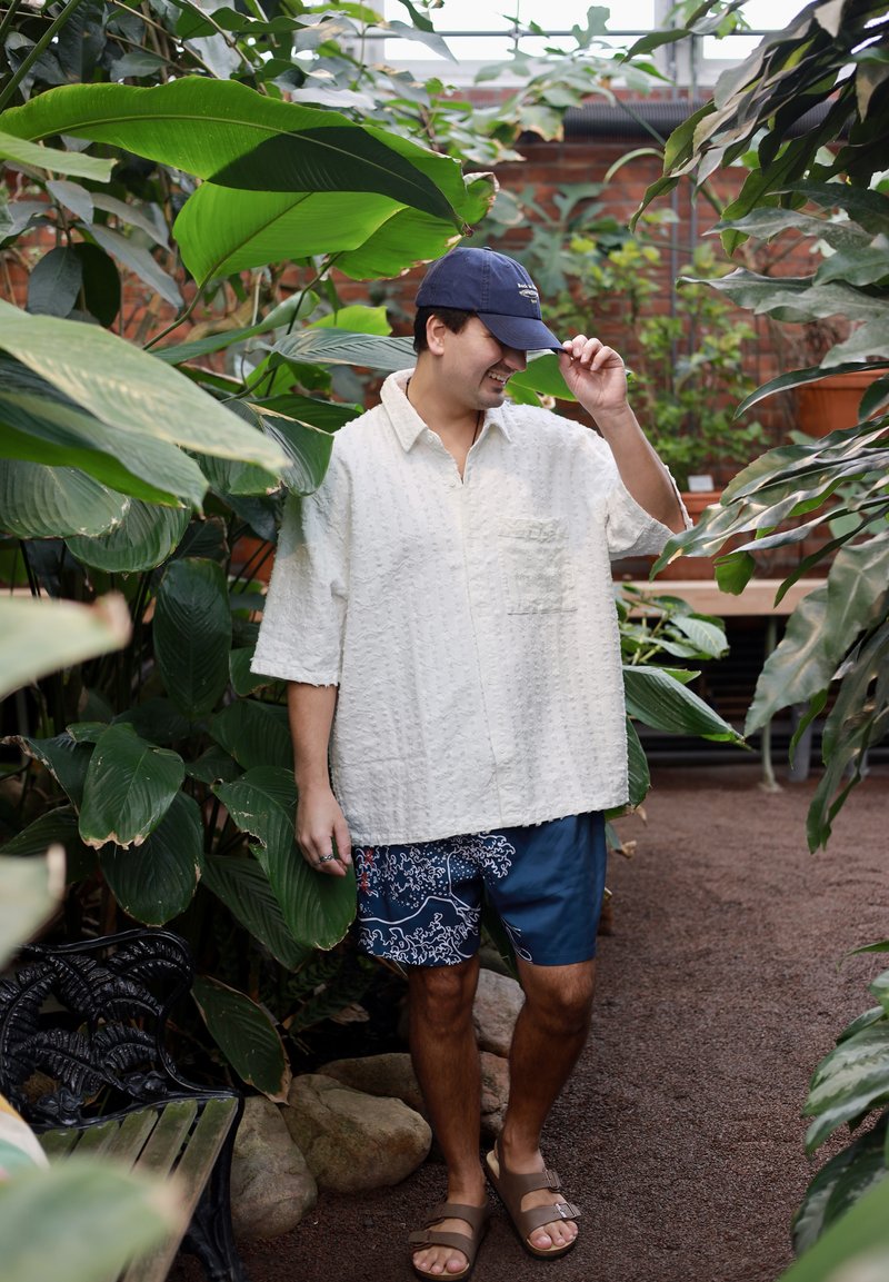 Mann, der ein weißes, strukturiertes Hemd, marineblaue, gemusterte Shorts, Sandalen und eine marineblaue Mütze trägt und zwischen großen grünen Pflanzen in Innenräumen steht.