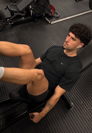 Hombre con ropa de entrenamiento negra usando la máquina de prensa de piernas en el gimnasio, expresión concentrada, con auriculares blancos.