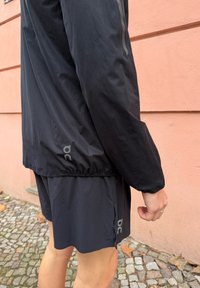 Zwart hardloopjack met een aansluitend ontwerp, elastische manchetten en een logodetail op de mouw. Gecombineerd met bijpassende shorts met zijopeningen.