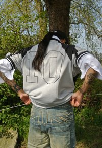 Graues Trikot mit schwarzen Ärmeln, mit der Nummer "10." Getragen über blauen Jeans mit auffälligen Nähten an den Hintertaschen. Hintergrund mit einem Baum.