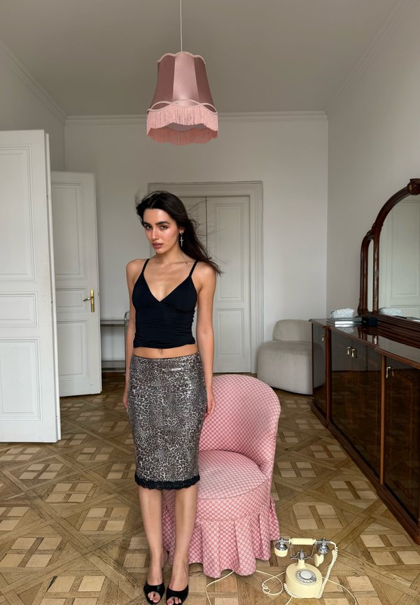 Junge Frau in schwarzem Top und Leopardenmuster-Rock steht neben einem pink gemusterten Stuhl mit einem Vintage-Drehtelefon auf Parkettboden.