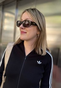 Schwarze gerippte Zip-Jacke mit weißen Streifen, mit einem kleinen weißen Adidas-Logo. Braune ovale Sonnenbrille und eine beige Umhängetasche sind sichtbar.