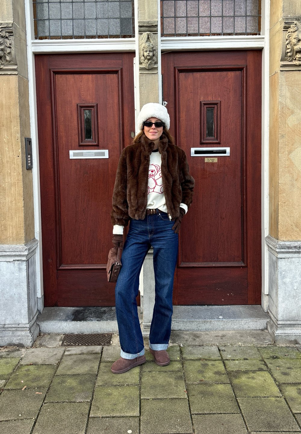 Casaco de pelúcia castanho, suéter creme com design vermelho, jeans azuis flare, luvas castanhas e botas cinzas. De pé em frente a portas de madeira.