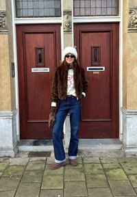 Casaco de pelúcia castanho, suéter creme com design vermelho, jeans azuis flare, luvas castanhas e botas cinzas. De pé em frente a portas de madeira.
