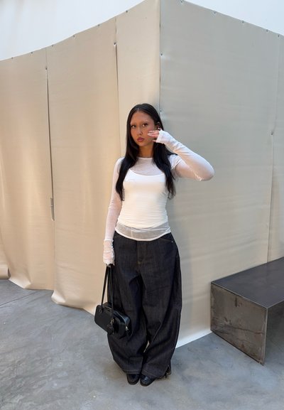 Mujer con cabello largo y oscuro, vestida con una blusa blanca de manga larga y pantalones anchos oscuros, sosteniendo un bolso negro, de pie en el interior junto a paredes beige.