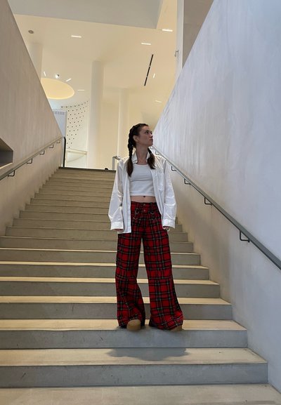 Camisa blanca extragrande, top corto blanco ajustado y pantalones anchos de cuadros rojos con bolsillos, de pie en una escalera gris en un entorno interior moderno.