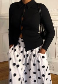 Top preto canelado de manga longa com decote de colarinho e detalhes em botão, combinado com uma saia branca com grandes manchas pretas.