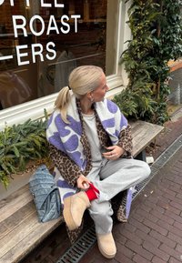 Leopardo e manta com padrão roxo, calças de fato cinzas, botas bege, bolsa acolchoada azul com alça de corrente e detalhe das meias vermelhas. Ambiente com banco de madeira.