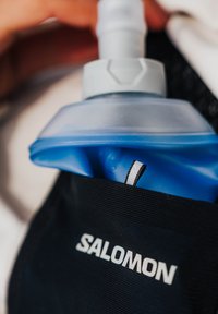 Main tenant une gourde de sport bleue partiellement insérée dans une pochette noire Salomon avec une languette réfléchissante.