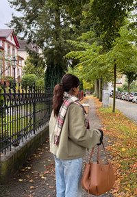 Übergroße hellgrüne Jacke, kariertes Tuch, hellblaue Jeans, trägt eine braune Lederhandtasche, umgeben von Herbstlaub und Gehweg.