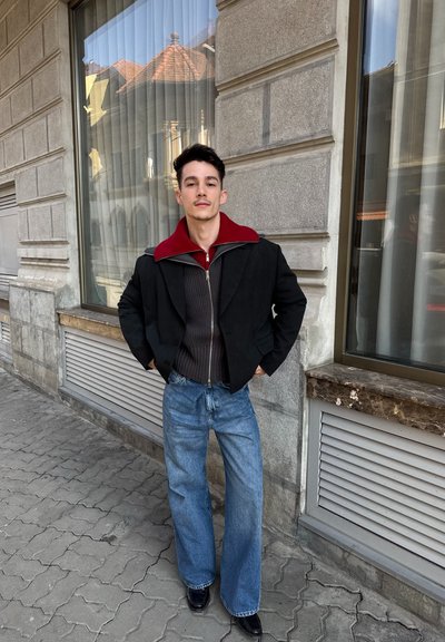 Joven con las manos en los bolsillos, de pie en la acera de la ciudad, vistiendo chaquetas de varias capas, vaqueros anchos azules y zapatos negros junto a un edificio de piedra.
