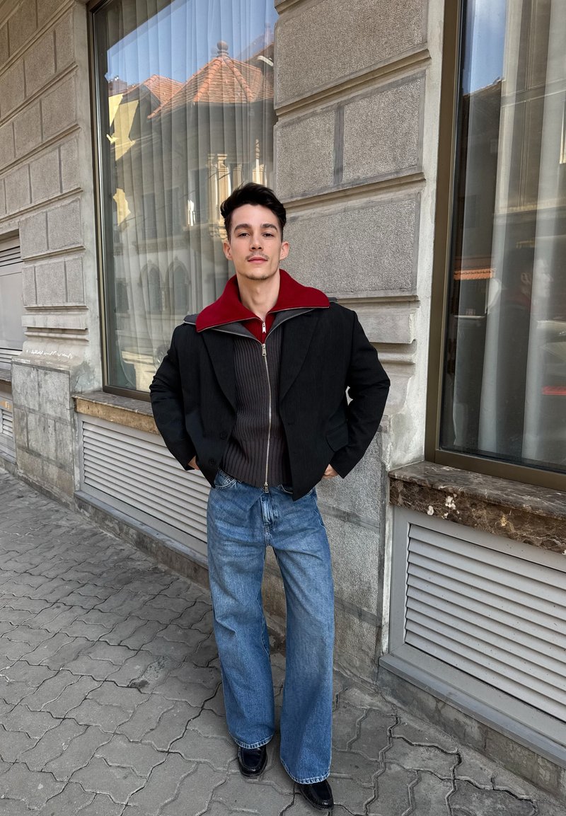 Νέος άνδρας με τα χέρια στις τσέπες στέκεται σε πεζοδρόμιο της πόλης φορώντας στρωτά μπουφάν, φαρδιά μπλε τζιν και μαύρα παπούτσια δίπλα σε πετρώδες κτίριο.