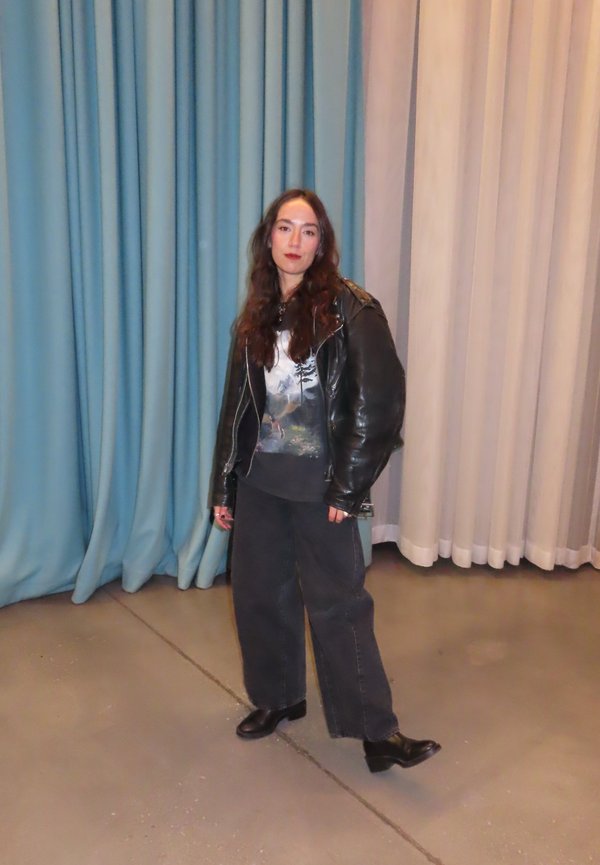 Black leather biker jacket over a graphic t-shirt, paired with wide black trousers and black ankle boots. Set against blue and beige drapes.