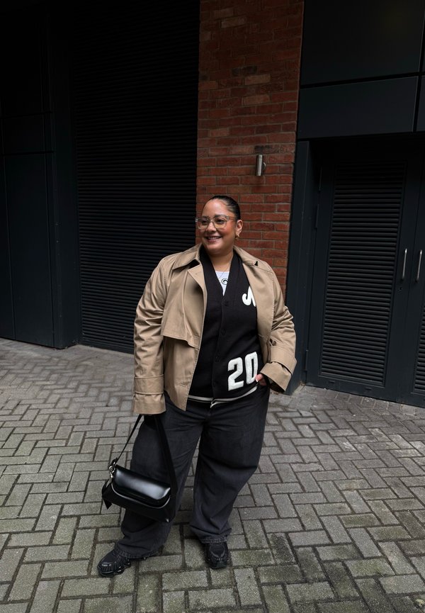 Persona con gafas, chaqueta beige, cárdigan negro con número blanco "20", pantalones negros, sosteniendo un bolso negro, de pie en una calle pavimentada junto a una pared de ladrillo.