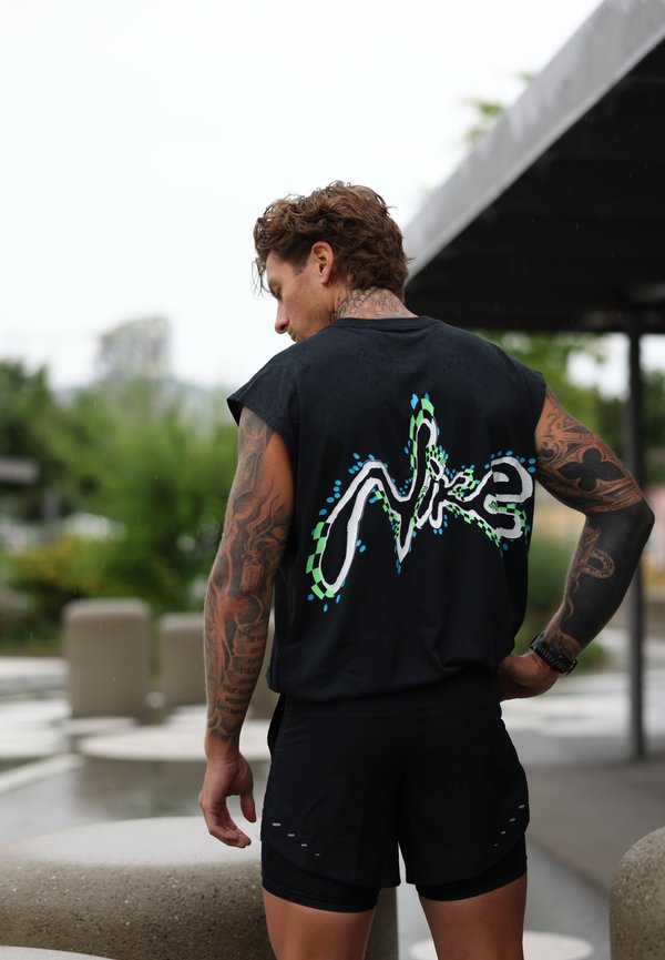 Homme tatoué porte un débardeur noir Nike avec un logo coloré dans le dos et un short noir, debout à l'extérieur, main sur la hanche.