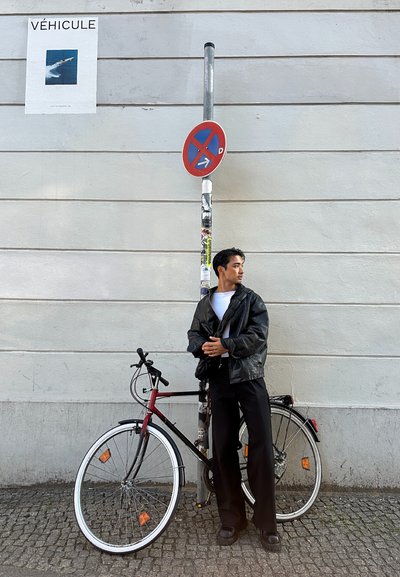 Una bicicleta roja y negra con una llanta blanca, aparcada contra un poste con un cartel de prohibido estacionar, al lado de una persona con una chaqueta y pantalones negros.