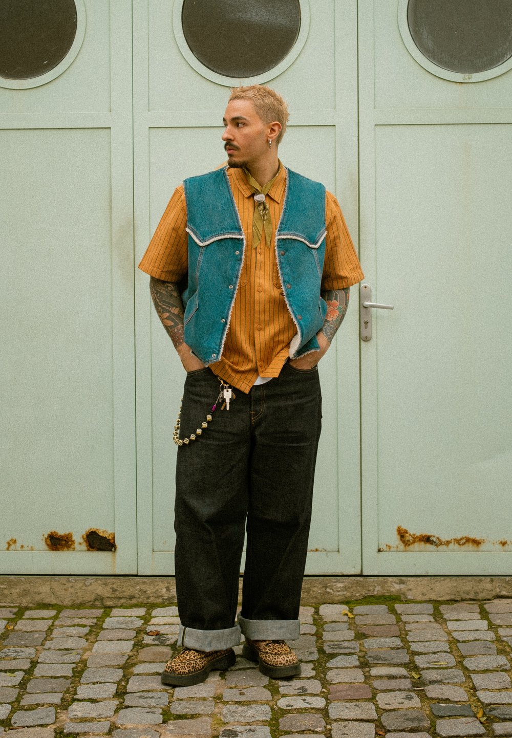 Veste en jean portée sur une chemise rayée orange, jeans larges noirs avec revers ; chaussures imprimé léopard et collier avec pendentifs. Fond gris.