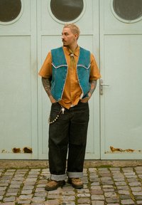 Veste en jean portée sur une chemise rayée orange, jeans larges noirs avec revers ; chaussures imprimé léopard et collier avec pendentifs. Fond gris.